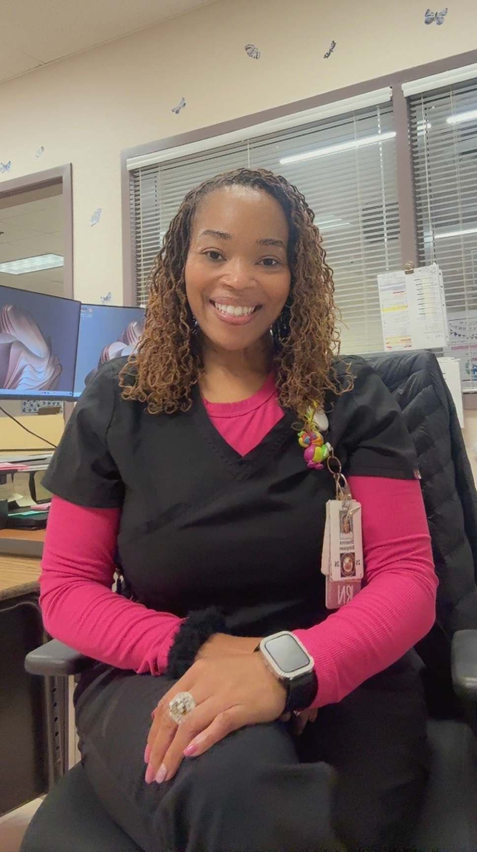 A smiling woman with curly hair wearing a black jacket and a pink top, sitting at a desk in an office environment.