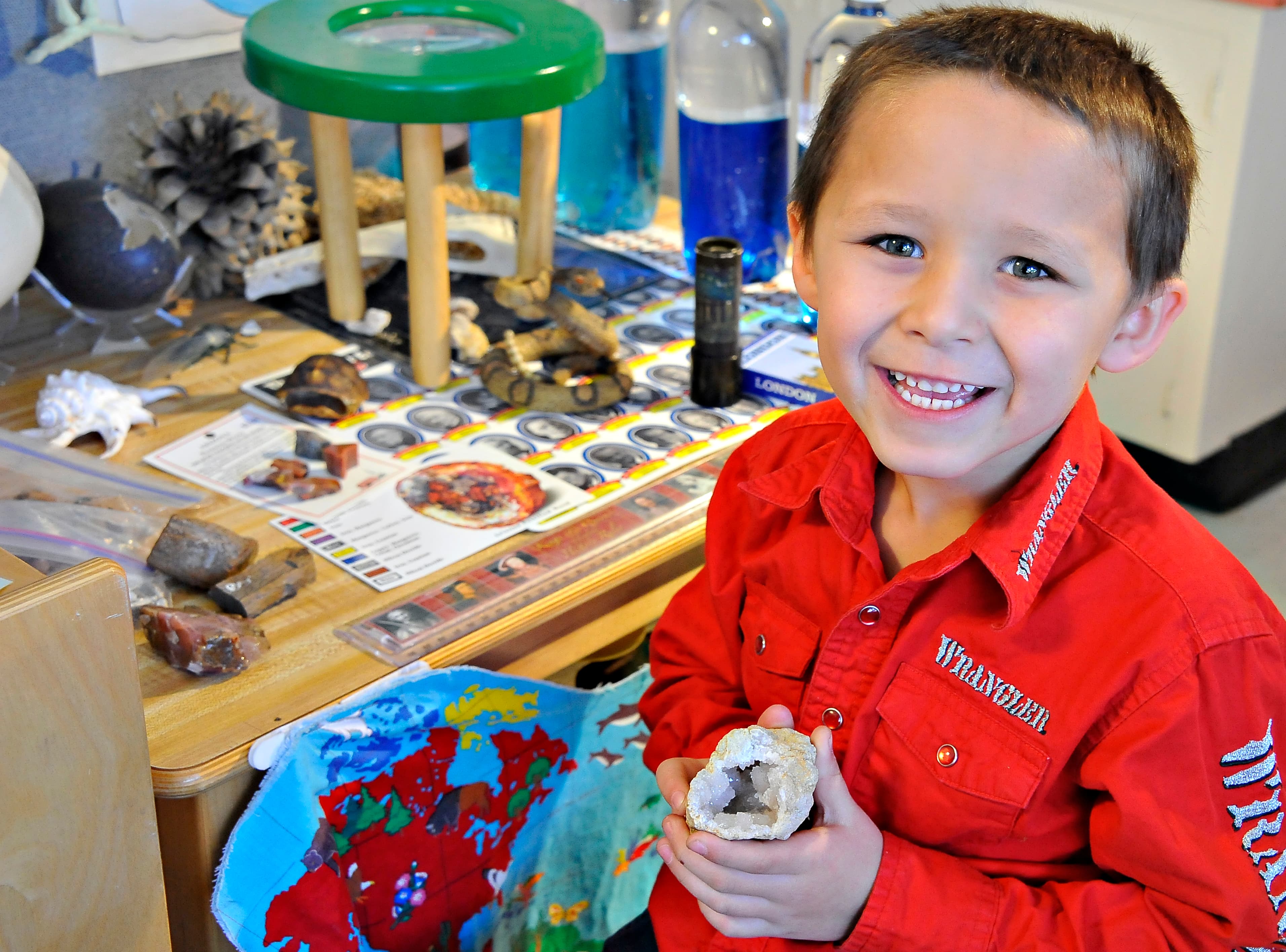 A little boy in a red shirt smiles and holds up a rock